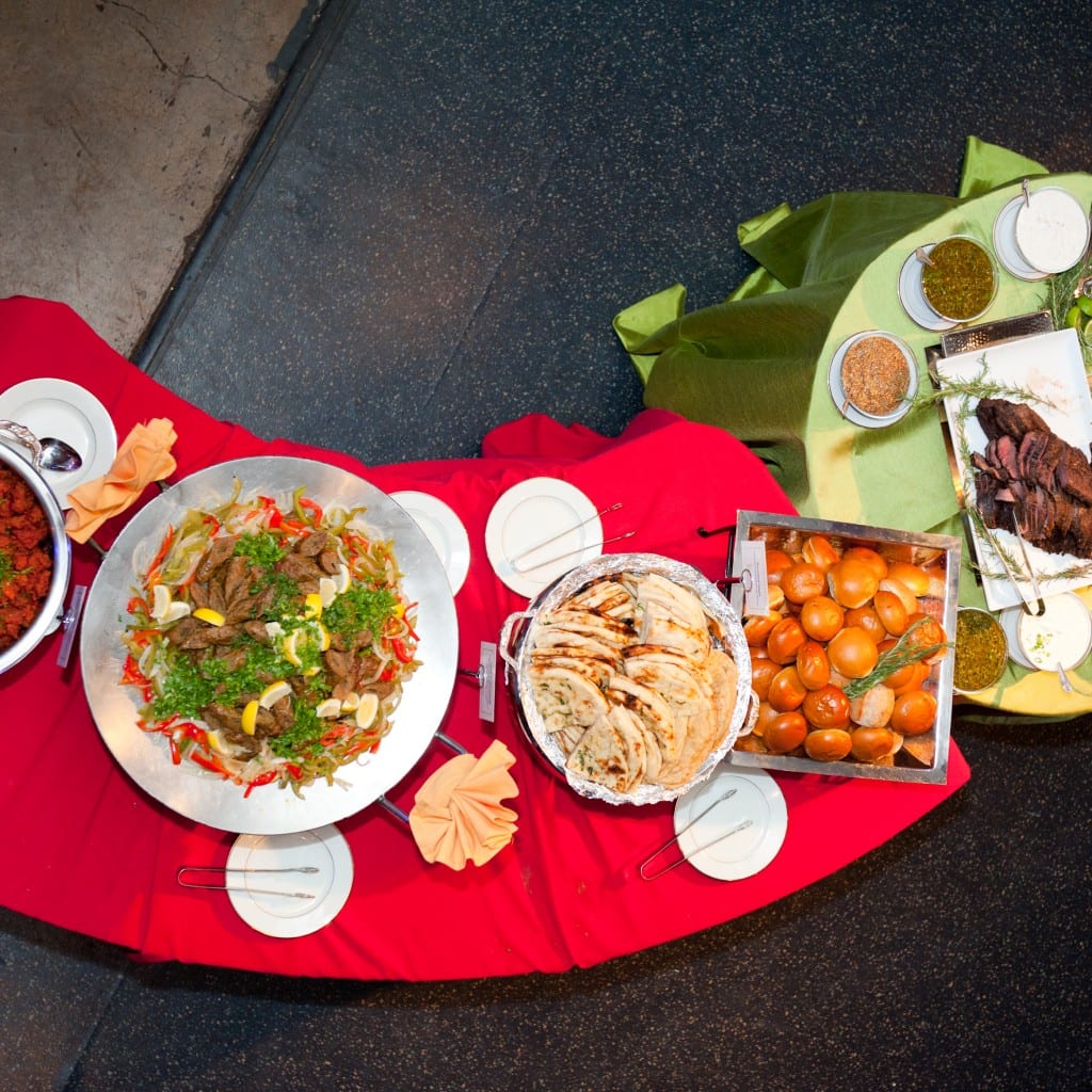 An IndAroma buffet dressed on linen and seen from above — a platter of grilled lamb, naan, pav, chutneys and raita with serving tongs and side plates.