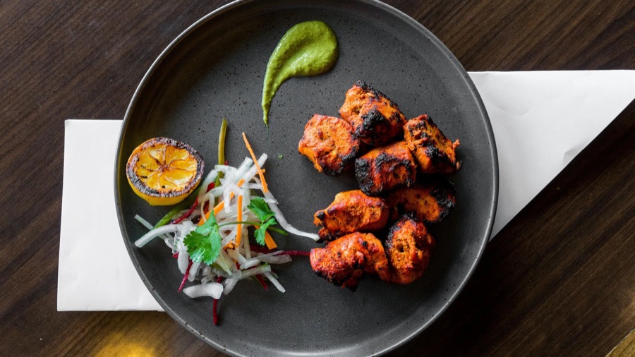 Charred chicken tikka on a dark plate with mint chutney, burnt lemon and a pickled salad.